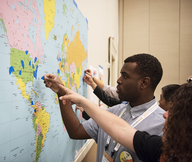 Young man stands in front of a map of the world taped on the wall, while woman points at a spot on the map